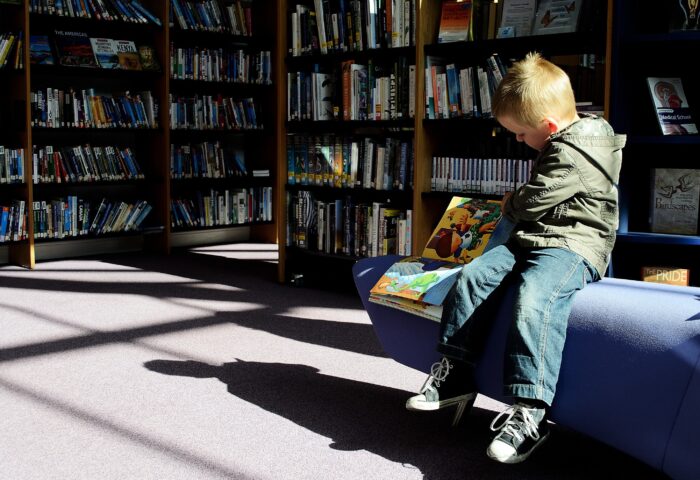 Young child sitting on bench in library scanning a book to his right side.+