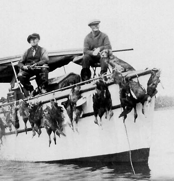 A photo of father and son returning on a boat from duck-hunting with a row of ducks straped to the stern of the boat,