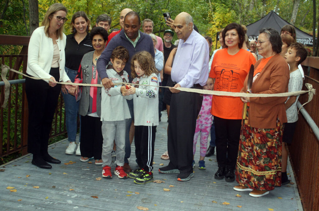 A large group of Manor Park residents and officials gathered Sept. 23 for the inauguration of the Sumac Footbridge, located at the eastern end of Blasdell Avenue. Cutting the ribbon to officially open the structure are Ismael Orion Di Carlo and Michael Acadia Di Carlo, whose father Anthony (standing behind) was a member of the Manor Park Community Council (MPCC) bridge naming committee. Also shown here (left to right) are Émilie Girard-Ruel of the National Capital Commission; Kelsey McMahon of the bridge naming committee; Mrs Saroj Aggarwal; Di Carlo: Rideau-Rockcliffe Councillor Rawlson King; Mr. Anand Aggarwal; Anita Tenasco and her mother Jenny Buckshot Tenasco of Kitigan Zibi. PHOTO: RYLAND COYNE